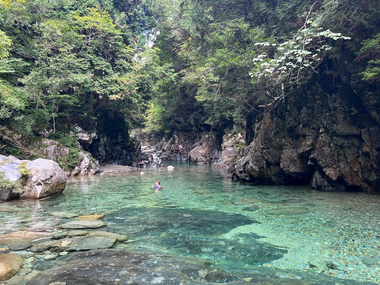 Swimmers in the emerald-green "Atera Blue" water of Atera Valley, framed by steep forested canyon walls